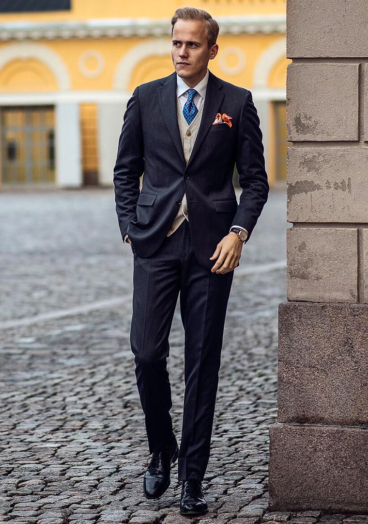 Black suit, white dress shirt, beige cardigan, blue tie, and black brogues