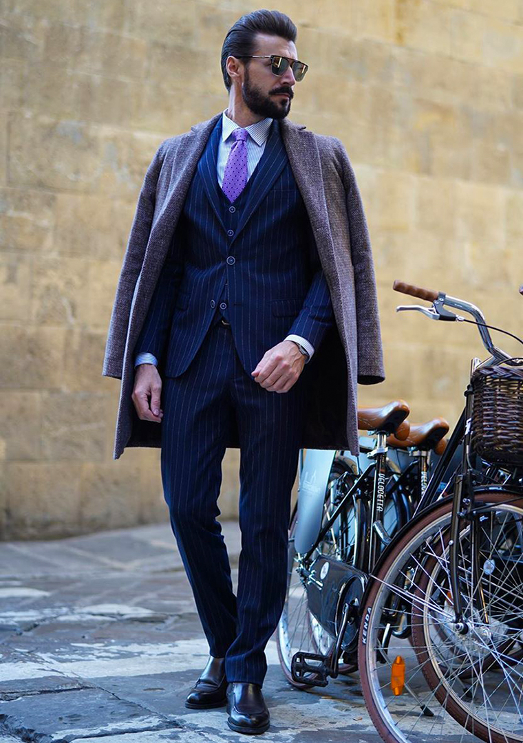 Navy three-piece pinstripe suit, grey overcoat, purple striped dress shirt, and black Chelsea boots